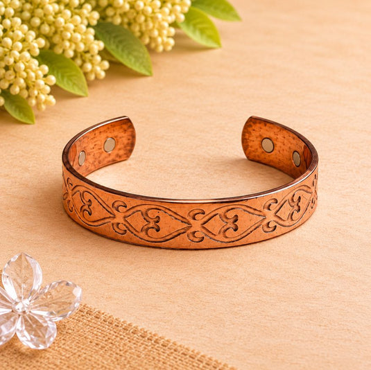 A copper-toned open cuff bracelet with engraved floral and scroll patterns, displayed on a beige surface with small decorative flowers in the corners.
