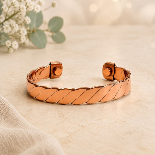 A copper-toned twisted cuff bracelet with a polished finish, displayed on a light beige textured surface with soft-focus flowers in the background.