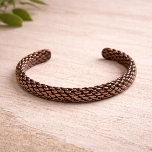Bronze braided bracelet on a light wooden surface with green leaves in the background