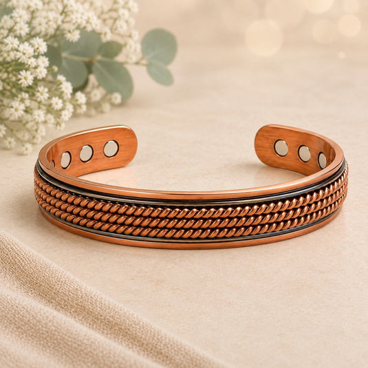 A copper-toned open cuff bracelet with a textured rope-like band and three circular inset details on each end, photographed on a soft beige surface with blurred white flowers in the background.