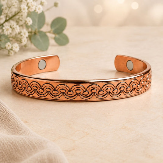 A copper-toned open cuff bracelet with an engraved circular wave pattern, featuring a small circular magnetic insert on each end, displayed on a light beige textured surface with soft-focus flowers in the background.