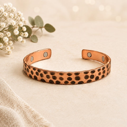 A hammered copper cuff bracelet with dark brown spots and small silver magnetic clasps, displayed on a beige fabric surface with dried flowers in the background.