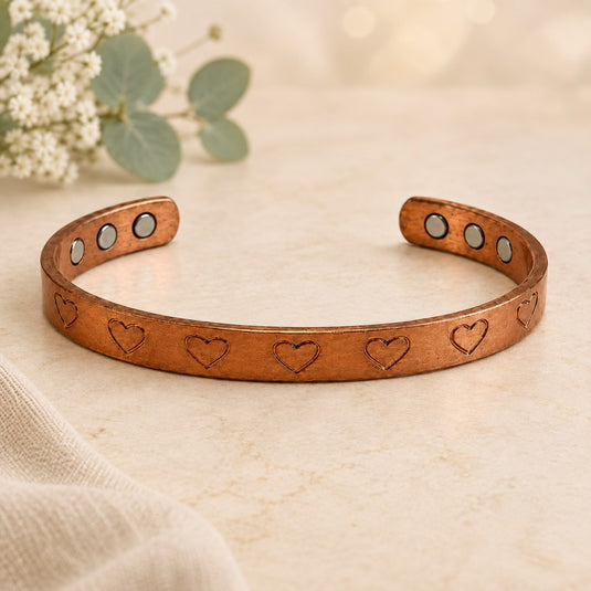 A copper-toned open cuff bracelet with engraved heart motifs and four circular magnetic inlays, displayed on a light beige textured surface with soft floral props in the background.