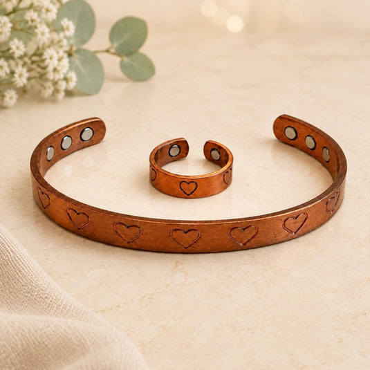 A copper-toned magnetic cuff bracelet and matching ring with engraved heart details, displayed on a light beige surface with soft floral props in the background.