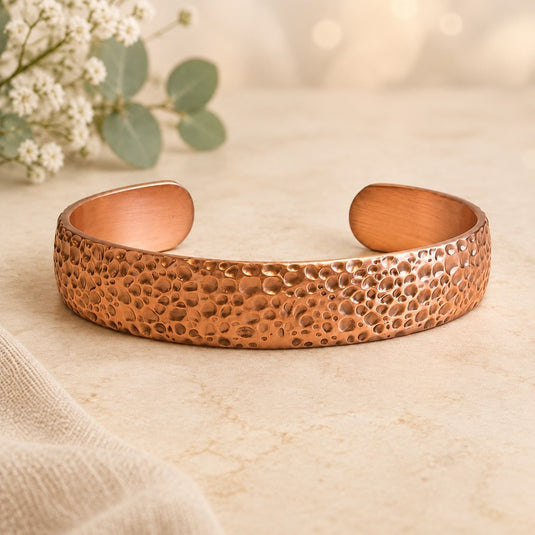 A hammered copper cuff bracelet with a textured, pebbled exterior and smooth, polished interior, displayed on a light beige stone surface with soft-focus eucalyptus and white flowers in the background.