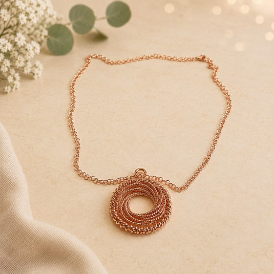 A rose gold necklace with a coiled, textured pendant, displayed on a warm beige textured surface with eucalyptus leaves and white flowers as props.