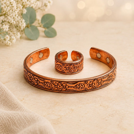 A copper-toned engraved cuff bracelet and matching ring displayed on a light beige surface with soft floral props in the background.