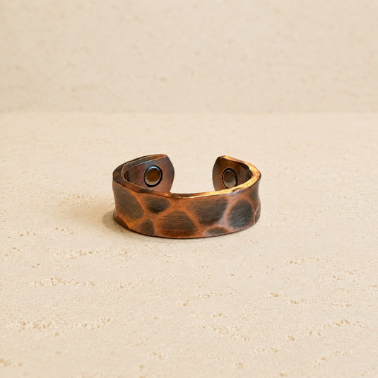 A hammered copper-toned metal cuff ring with a textured, organic pattern of dark and light brown patches, resting on a light beige textured surface.