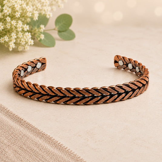 A braided cuff bracelet in a warm copper or bronze tone with small white inlay details, photographed on a soft beige surface with blurred flowers in the background.