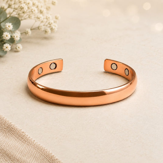 A polished rose gold cuff bracelet with two circular magnetic therapy inserts on each end, displayed on a textured beige surface with small white flowers in the background.
