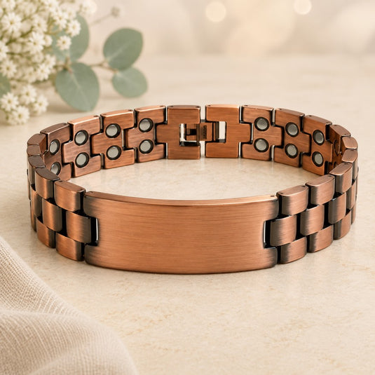 A gold metal link bracelet with a brushed central plate, displayed on a light beige textured surface with soft-focus eucalyptus leaves in the background.