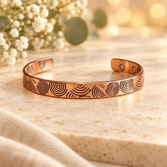 A copper-colored open cuff bracelet with engraved spiral and wave patterns, displayed on a light stone surface with soft floral props and warm bokeh in the background.