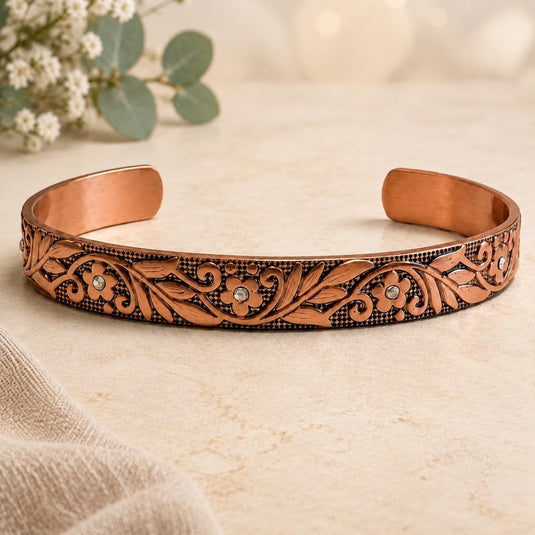 An open cuff bracelet made of copper with an engraved floral and scroll pattern, accented with small clear rhinestones, resting on a light beige textured surface with soft-focus flowers in the background.