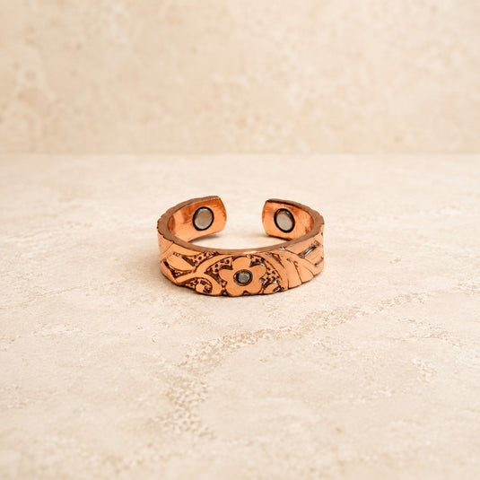 A handmade copper cuff bracelet with engraved floral patterns and two circular black and white stone accents, displayed on a textured beige surface.