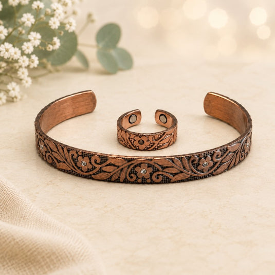 A copper-toned open cuff bracelet and matching adjustable ring, both engraved with floral and scroll motifs and accented with small clear stones, displayed on a light beige surface with soft blurred flowers in the background.