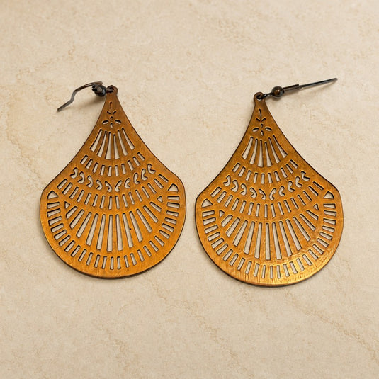 A pair of teardrop-shaped dangling earrings with intricate cutout patterns, made from a warm gold-toned metal, displayed on a textured beige surface.