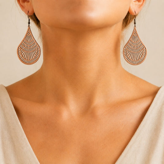 Close-up of a woman wearing large teardrop-shaped dangling earrings with an intricate openwork pattern in a warm metallic copper tone against a soft beige background.
