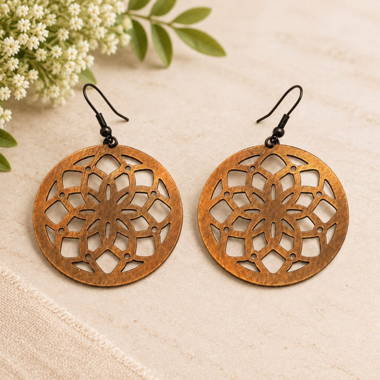 A pair of large circular copper-toned dangle earrings with intricate cutout floral patterns and black fishhook wires, displayed on a light beige textured surface with small white flowers and green leaves in the corner.