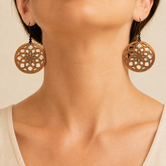 Close-up of a person wearing large circular wooden dangle earrings with an intricate cutout floral pattern, against a warm beige background.