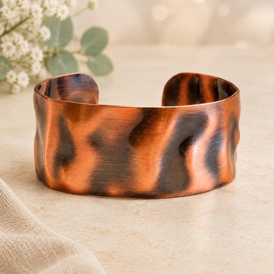 A hammered copper cuff bracelet with dark oxidized accents, displayed on a light beige surface with soft-focus flowers in the background.