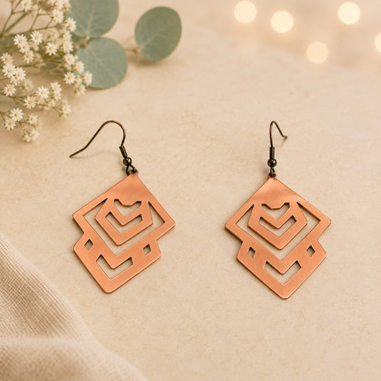 A pair of geometric drop earrings with a copper-toned cutout design and black fishhook wires, displayed on a beige textured surface with decorative flowers and soft bokeh lights.