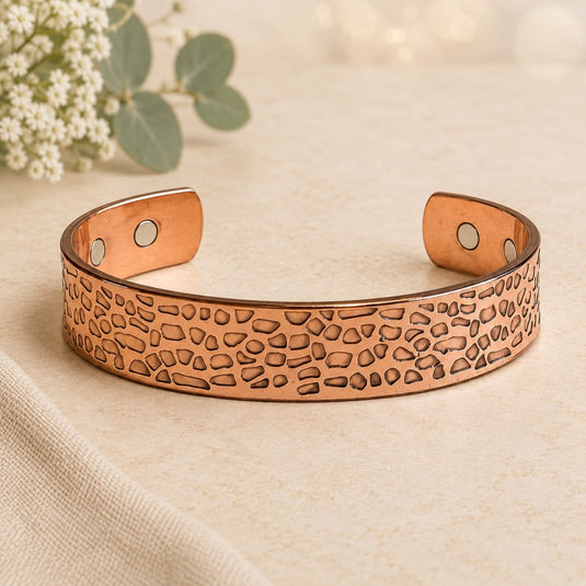A copper-toned open cuff bracelet with a textured, hammered stone-like pattern and two small circular magnets on the inner ends, displayed on a light beige fabric surface with soft floral props in the background.