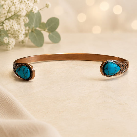 A copper-toned open cuff bracelet with two oval turquoise stone settings, displayed on a light beige surface with soft bokeh lights and small white flowers in the background.