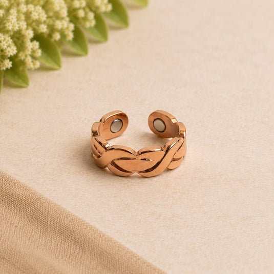 A rose gold adjustable ring with an interlocking braided design, resting on a textured beige surface with a soft-focus background of green leaves and white flowers.