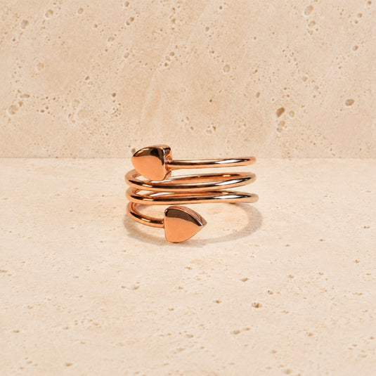 A rose gold wrap ring with two small heart-shaped charms, displayed on a textured beige stone surface.