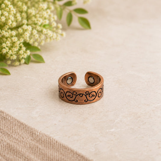 A copper-toned open cuff ring with black scrollwork engraving and circular magnetic stone inserts, displayed on a beige stone surface with small white flowers in the background.