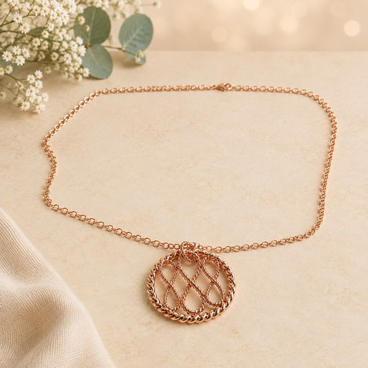 A rose gold chain necklace with a circular pendant featuring a woven lattice pattern, displayed on a textured beige surface with eucalyptus leaves and white flowers in the background.
