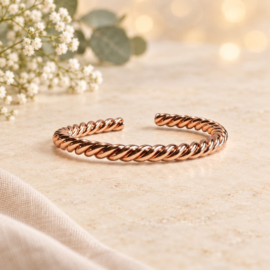A twisted rope-style cuff bracelet in polished rose gold, displayed on a light beige marble surface with soft-focus white flowers and warm bokeh lights in the background.