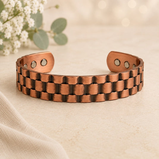 A copper-toned magnetic cuff bracelet with a textured, interlocking link design, displayed on a neutral beige fabric surface with soft white flowers in the background.