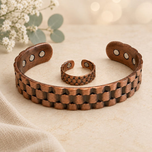 A copper-toned magnetic therapy bracelet and matching ring with a textured, interlocking link design, displayed on a neutral beige fabric surface with soft white flowers in the background.