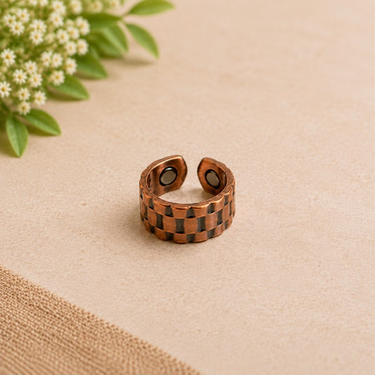 A copper-toned adjustable ring with a checkered pattern of dark brown and black squares, displayed on a beige textured surface with small white flowers in the corner.