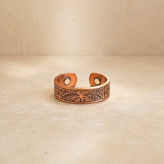 A copper-toned adjustable ring with an engraved tree design and circular magnetic snap closures, displayed on a textured beige surface.