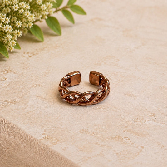 A copper-toned metal cuff ring with a twisted, braided design and square end caps, resting on a textured beige stone surface with small white flowers in the top-left corner.