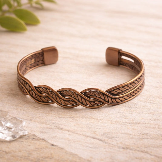 Bronze braided bracelet on a light wooden surface with green leaves in the corner.