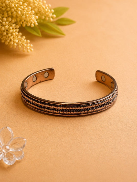 A copper-toned open cuff bracelet with a braided textured center stripe and three circular magnetic snap buttons on the inner band, displayed on a warm beige background with decorative yellow flowers in the corner.