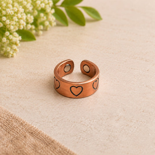 A copper-toned adjustable ring with engraved heart motifs, displayed on a textured beige surface with green leaves and small white flowers in the background.