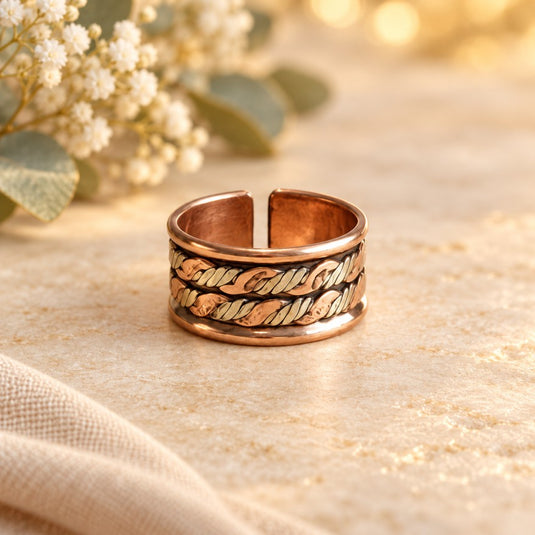 A copper-toned open cuff ring with a textured, hammered leaf pattern, displayed on a light beige textured surface with soft-focus baby's breath flowers in the background.
