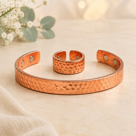 A copper-toned hammered metal cuff bracelet with a matching smaller matching ring, displayed on a light beige surface with soft floral props in the background.