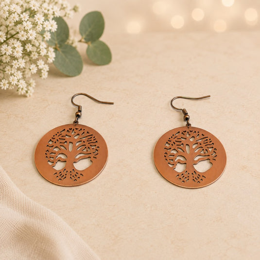 A pair of round copper-toned tree-of-life cutout dangle earrings with dark metal fishhook wires, displayed on a light beige textured surface with small white flowers and green leaves in the corner.