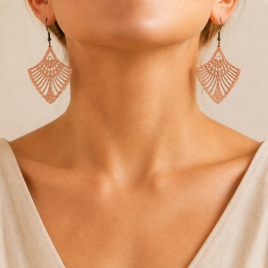 Close-up of a woman's neck and lower face wearing a pair of dangling fan-shaped earrings with an intricate openwork lace-like pattern in a warm metallic pink tone.