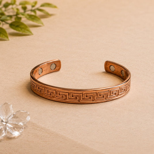 Copper bracelet with Greek key design on a beige surface with a plant and crystal in the background