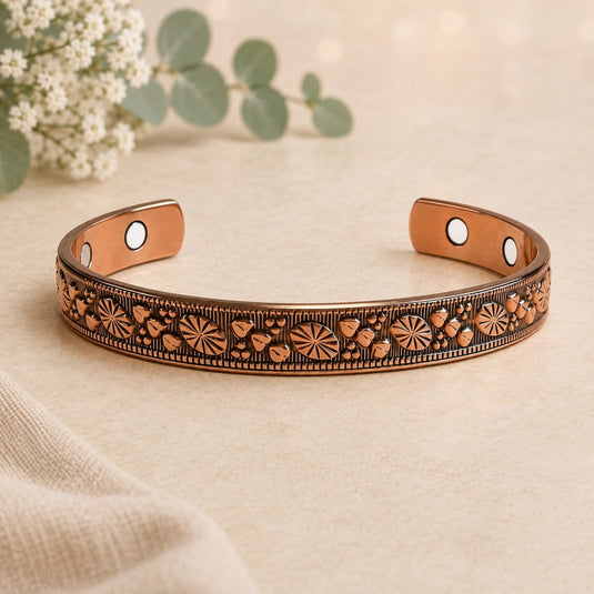 A copper-toned open cuff bracelet with engraved floral and geometric patterns, displayed on a beige fabric surface with soft-focus eucalyptus leaves and white flowers in the background.