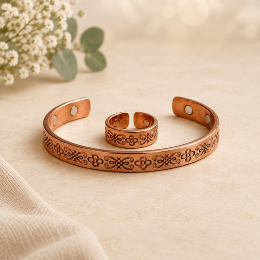 A copper-toned open cuff bracelet with engraved floral patterns, shown alongside a matching adjustable ring on a soft beige surface with blurred flowers in the background.