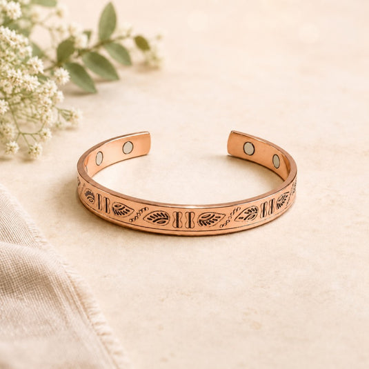 A copper-toned open cuff bracelet with engraved leaf and stripe patterns, photographed on a light beige surface with soft floral props in the background.