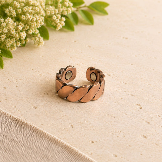 A copper-toned open cuff ring with a textured, leaf-like segmented design, resting on a light beige textured surface with green leaves and small white flowers in the top-left corner.
