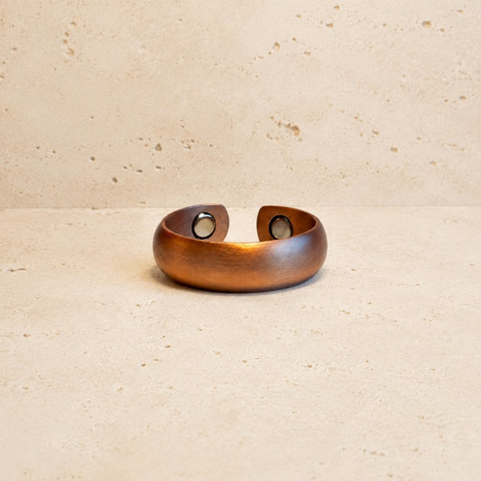 A hammered copper-toned metal cuff bracelet with two circular magnetic closures, displayed on a textured beige stone surface.